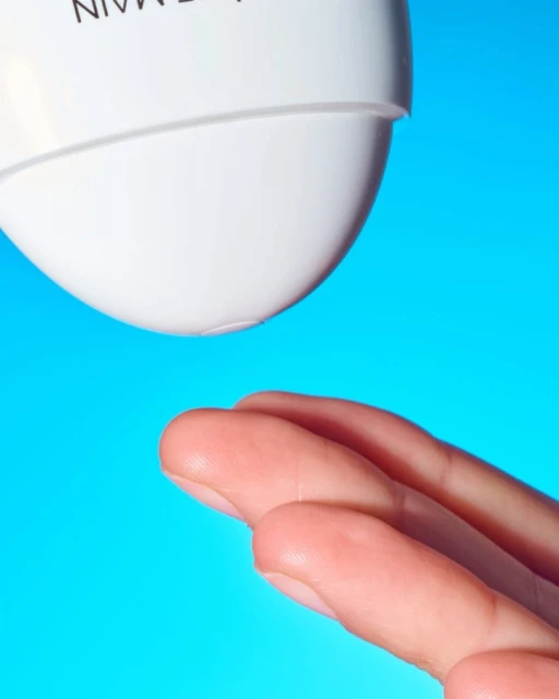 Close-up studio shot of fingers positioned under the nozzle of a white bottle against a vibrant blue background, suggesting skincare or beauty.