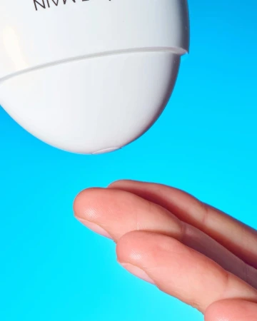 Close-up studio shot of fingers positioned under the nozzle of a white bottle against a vibrant blue background, suggesting skincare or beauty.