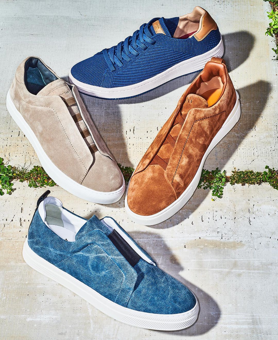 Close-up studio shot of three pairs of casual shoes in blue, tan, and brown leather, displayed on a textured stone surface.