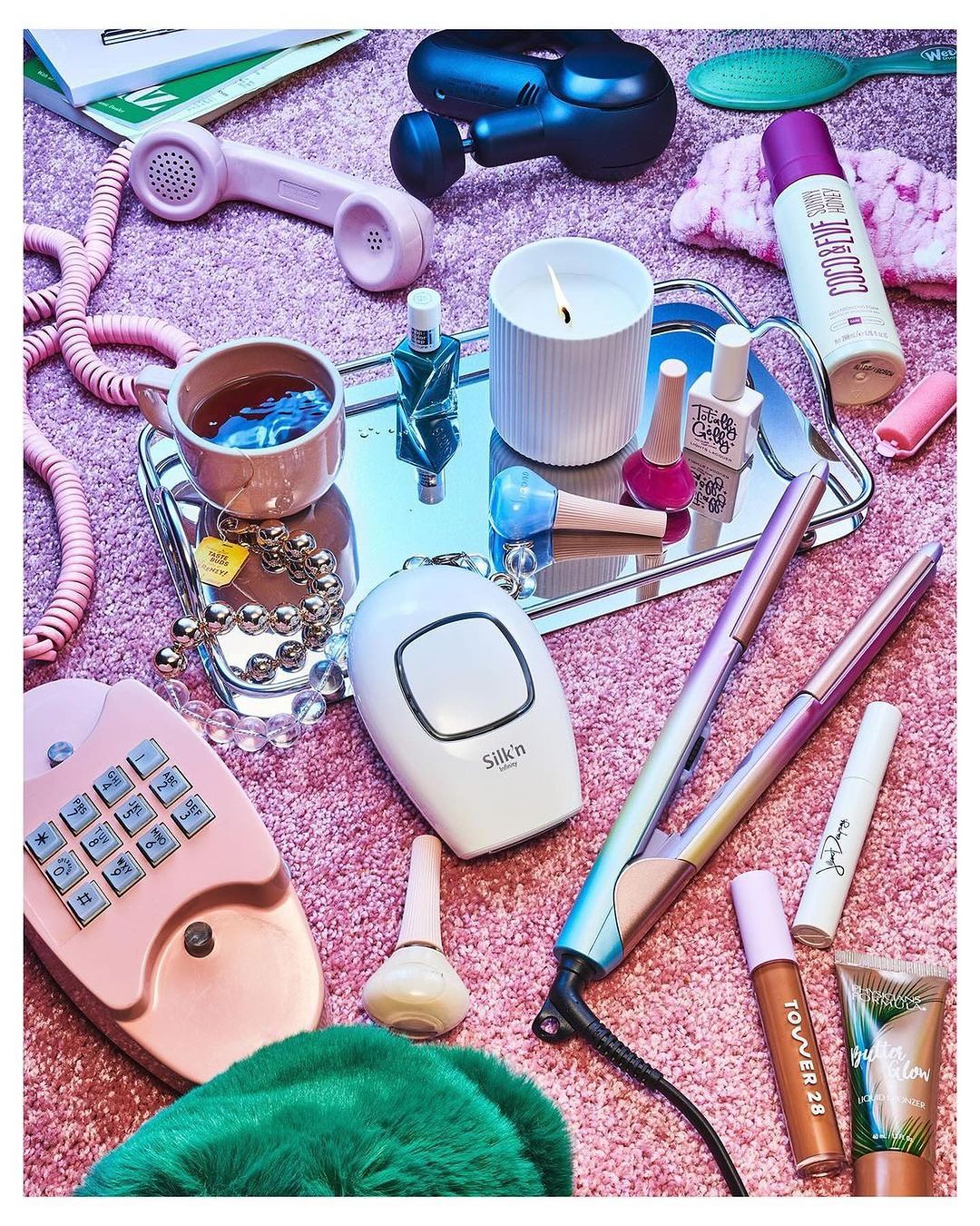 Still life of beauty and self-care products on a pink furry surface, including cosmetics, hair styling tools, and personal care devices.
