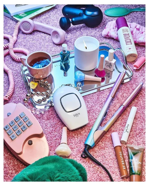 Still life of beauty and self-care products on a pink furry surface, including cosmetics, hair styling tools, and personal care devices.