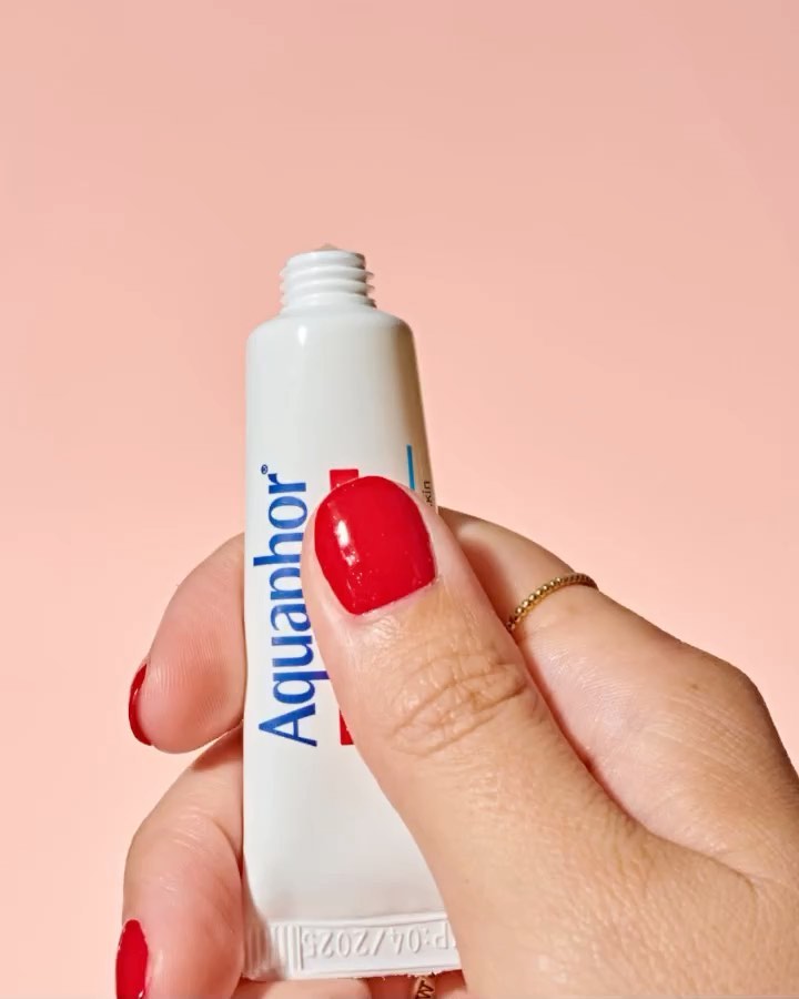Close-up shot of a hand holding a white tube of Aquaphor. The hand has red nail polish and a simple gold ring on one finger.