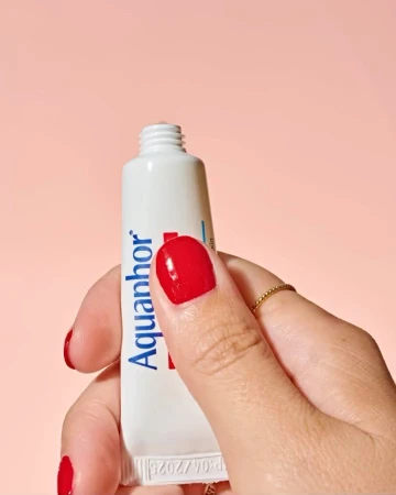 Close-up shot of a hand holding a white tube of Aquaphor. The hand has red nail polish and a simple gold ring on one finger.