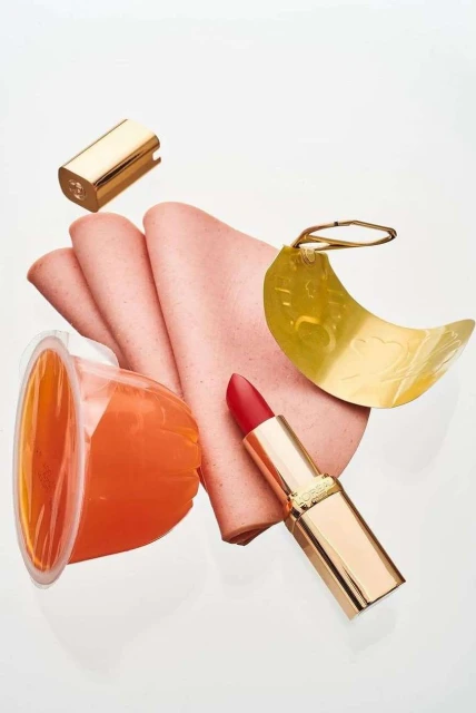 Still life of lipstick, lunch meat, orange gelatin cup, and gold-colored items against a white backdrop in portrait orientation.
