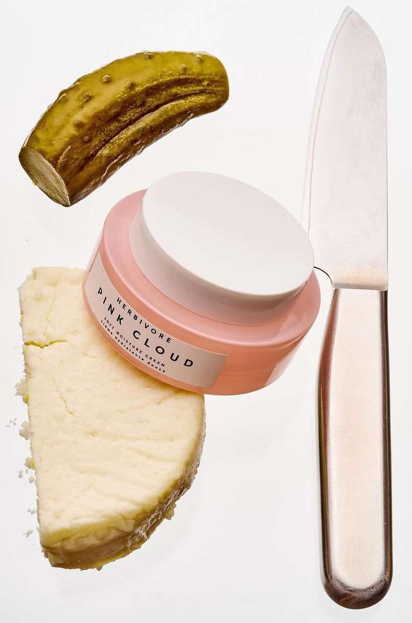 Cosmetic cream, cheese, knife, and pickle arranged in a still life composition against a white background, shot in studio.