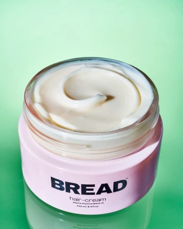 Open jar of off-white hair cream with a pink container against a solid green background