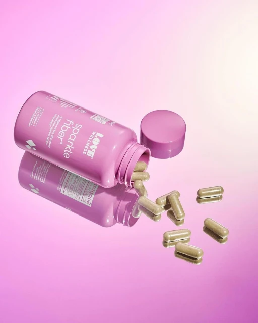 Pink bottle of Love Wellness Sparkle Fiber supplements with scattered capsules on a pink gradient reflective surface.