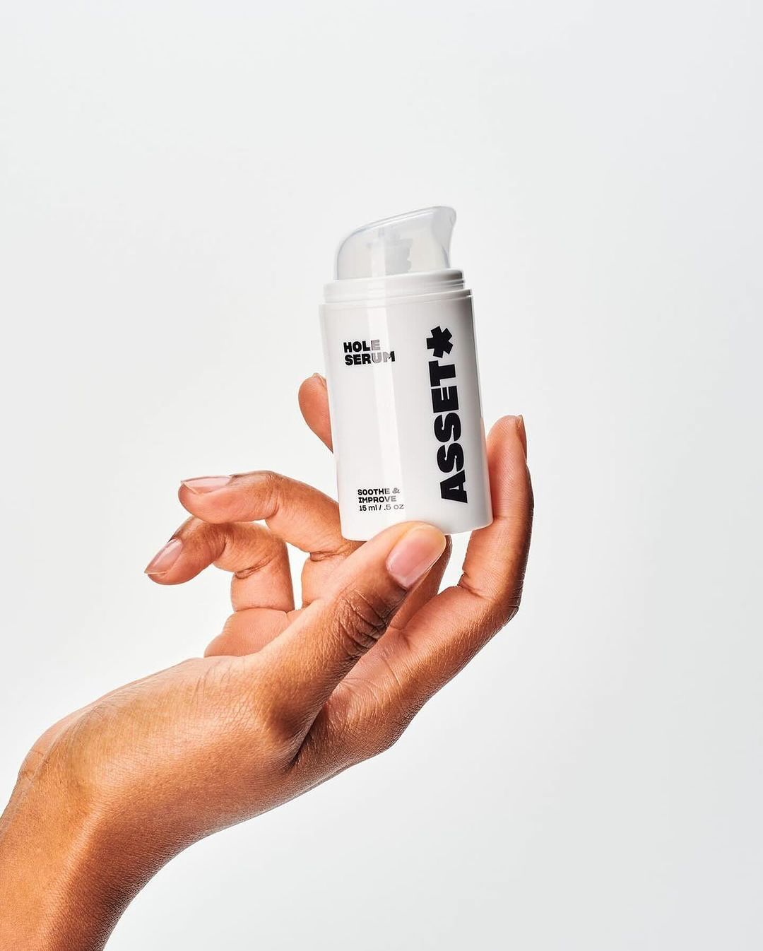 A woman's hand holds a white bottle of Asset Hole Serum against a bright background, showcasing the product in a beauty context.