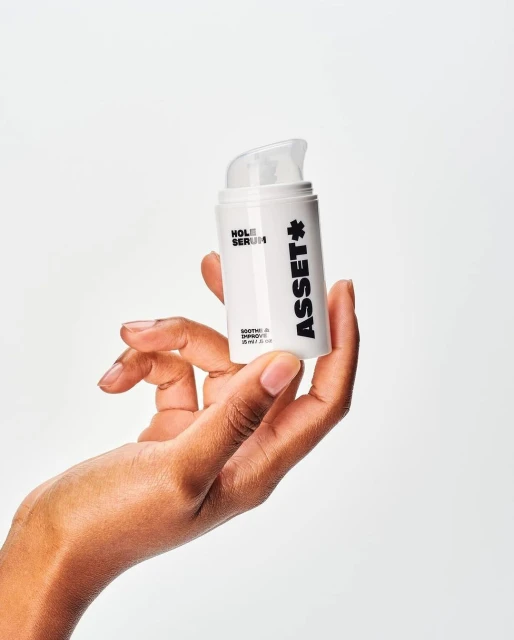 A woman's hand holds a white bottle of Asset Hole Serum against a bright background, showcasing the product in a beauty context.