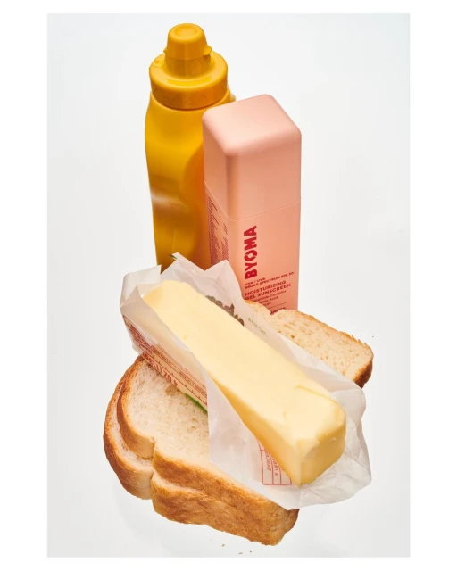 Still life photograph featuring a bottle of mustard, moisturizer, block of butter and slices of bread on a bright white background.