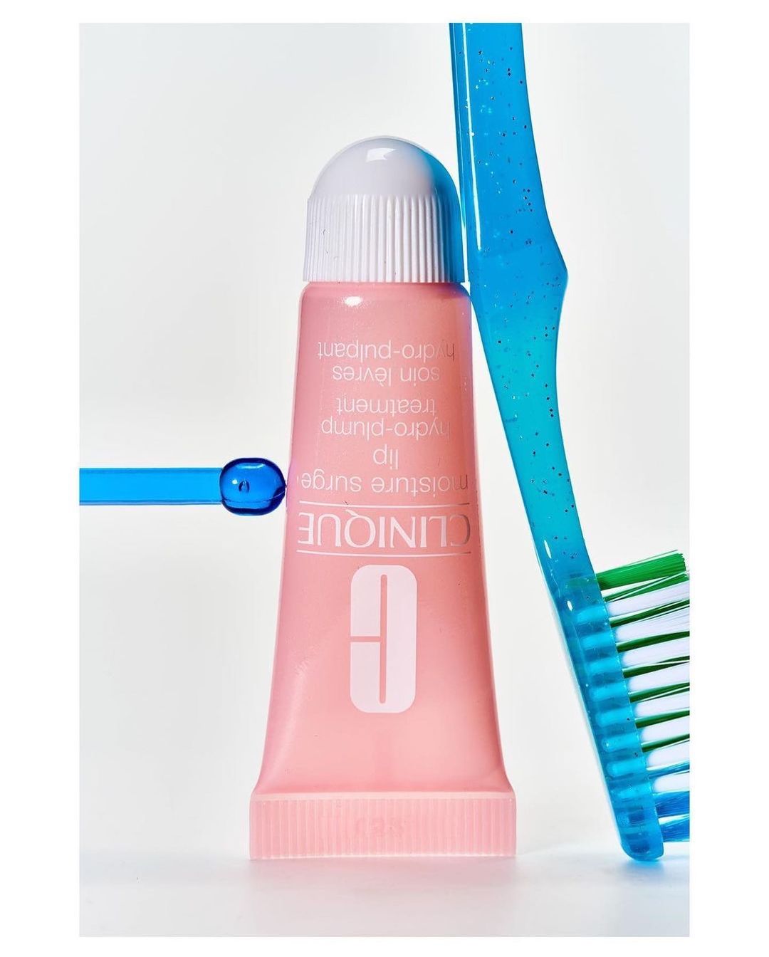 Close-up shot of a Clinique lip moisture surge treatment tube beside a blue and green toothbrush on a white background.