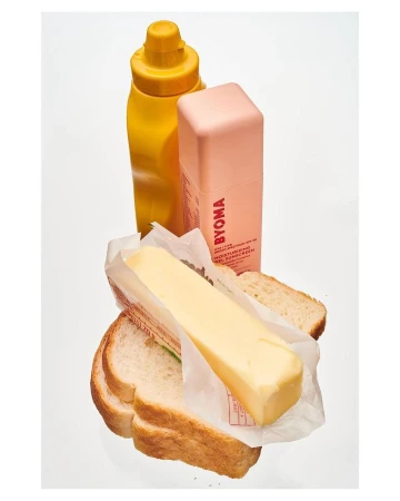 Still life of a bottle of mustard, a tube of moisturizing sunscreen, and a stick of butter on slices of white bread. 
