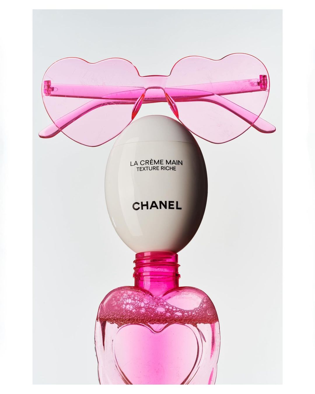 Still life shot of Chanel La Crème Main and pink heart-shaped sunglasses against a white backdrop, conveying beauty and luxury.