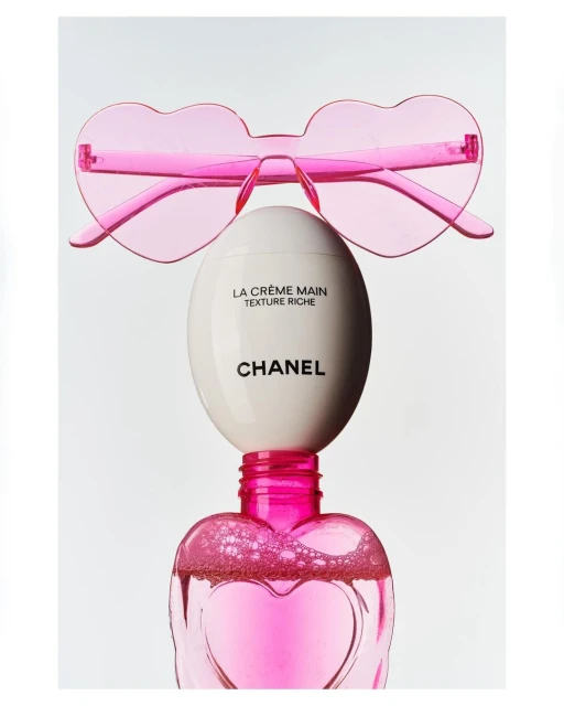 Still life shot of Chanel La Crème Main and pink heart-shaped sunglasses against a white backdrop, conveying beauty and luxury.