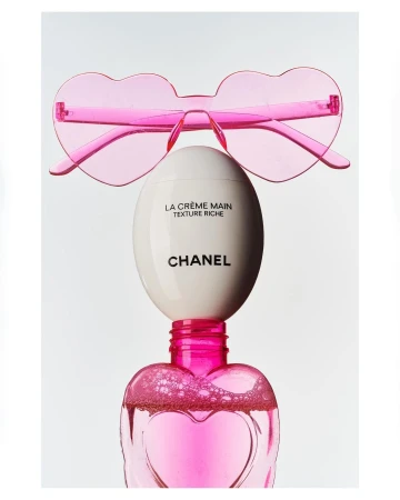 Still life shot of Chanel La Crème Main and pink heart-shaped sunglasses against a white backdrop, conveying beauty and luxury.