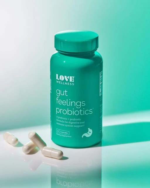 A green bottle of probiotics is on a white surface, along with three white probiotic capsules, against a gradient green background.