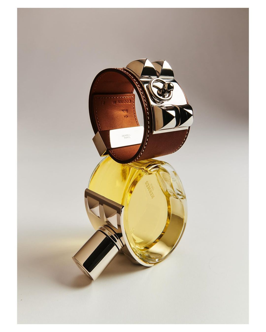 Still life featuring a brown leather Hermès bracelet with silver accents and a yellow Hermès perfume bottle, set against a neutral background.