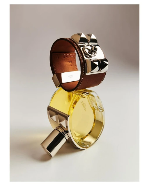 Still life featuring a brown leather Hermès bracelet with silver accents and a yellow Hermès perfume bottle, set against a neutral background.