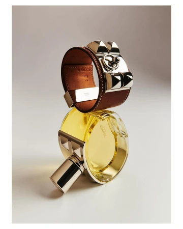 Hermes leather cuff bracelet resting on top of a round glass perfume bottle against a neutral gradient background.