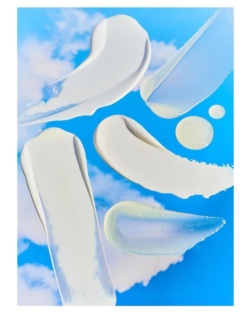 Cream smears on a bright sky-blue background with white clouds. Various sizes and shapes of cosmetic product swatches, shot in studio lighting.