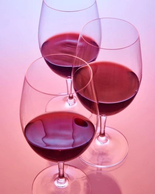 Three wine glasses filled with red wine are arranged on a pink surface, creating a visually appealing composition.