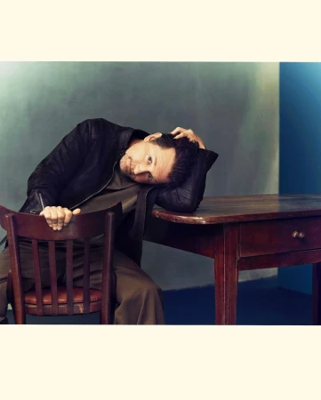 A man seated at a wooden table resting his head on his hand by photographer Joe Pugliese