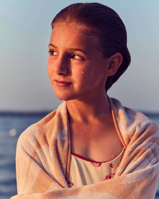 Young girl wrapped in a towel stands near the sea. She has short brown hair and a thoughtful expression while looking off into the distance.