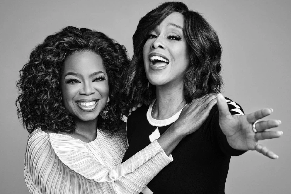 Black and white portrait of Oprah Winfrey and Gayle King smiling and embracing in a photo studio with a solid gray background.