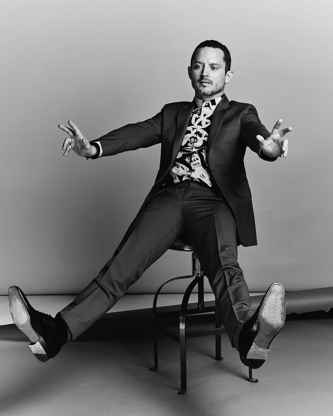 Monochrome portrait of a man in a tailored suit sitting on a metal stool with arms outstretched.
