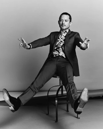 Monochrome portrait of a man in a tailored suit sitting on a metal stool with arms outstretched.