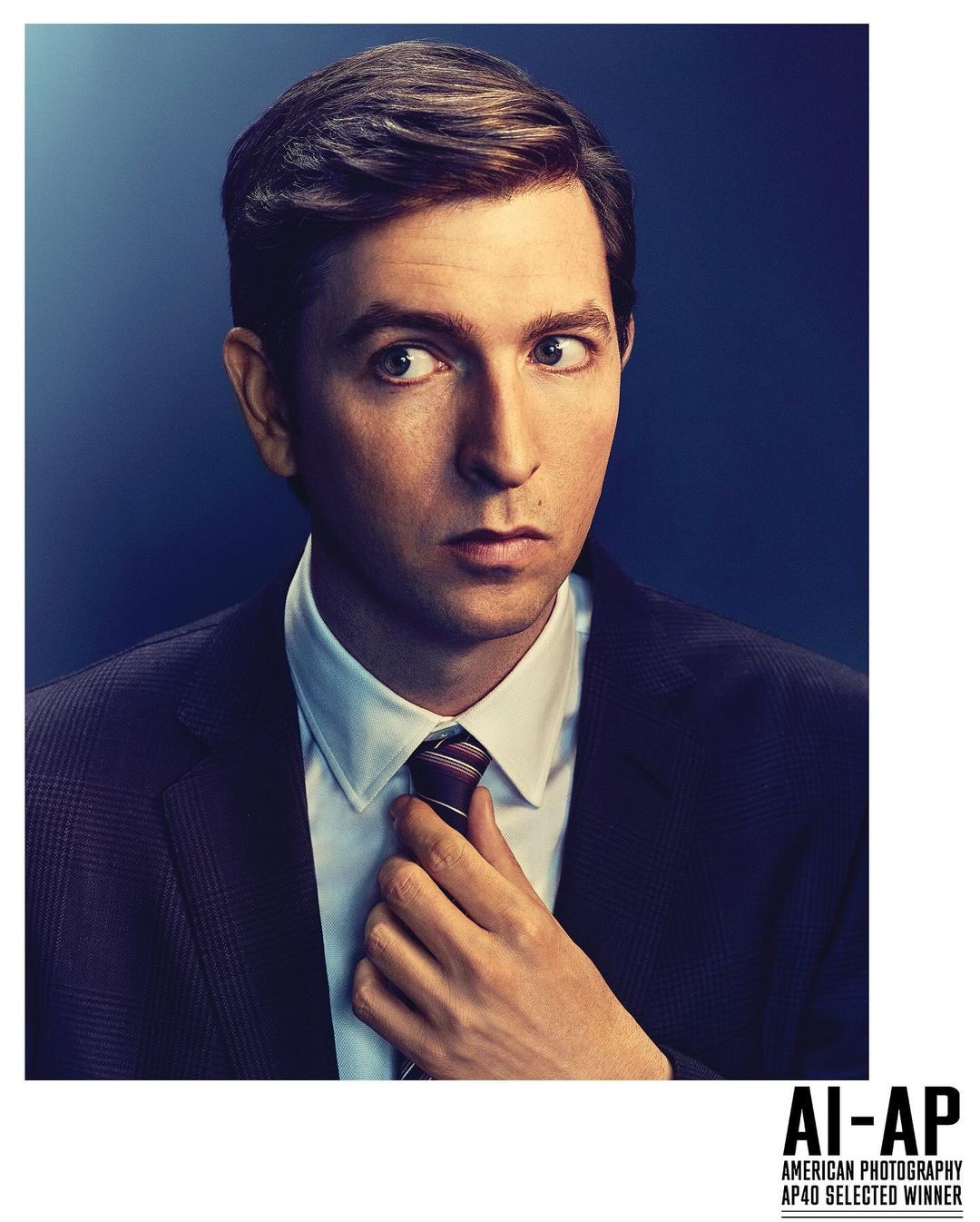 Studio portrait of a man in a suit adjusting his tie, looking sideways with a slightly apprehensive expression against a dark blue backdrop.