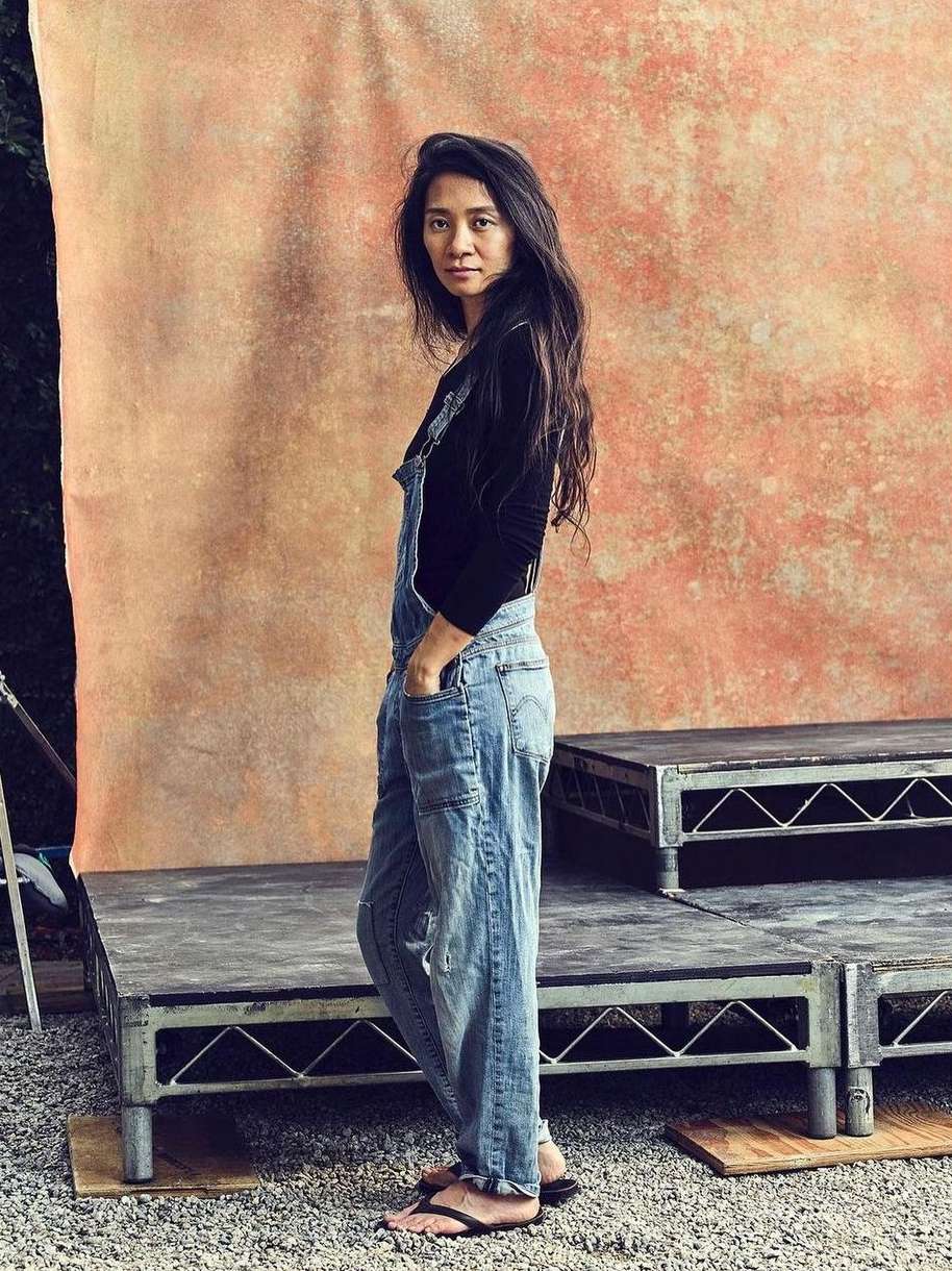 Portrait of an Asian adult female in denim overalls standing on a platform with a faded background in an outdoor setting.