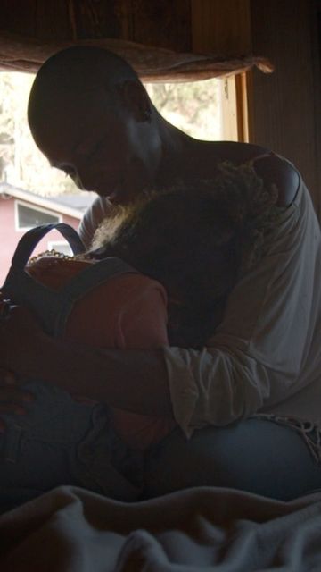 A softly lit indoor shot features a close embrace between a woman and a bald adult male, suggesting a tender or familial connection.