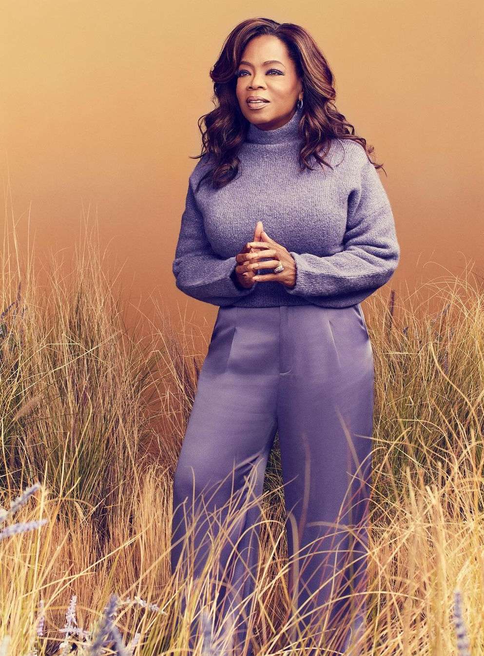 Oprah Winfrey posing in a sweater and pants in a wheat field, looking left, against a gradient orange background in a portrait shot.