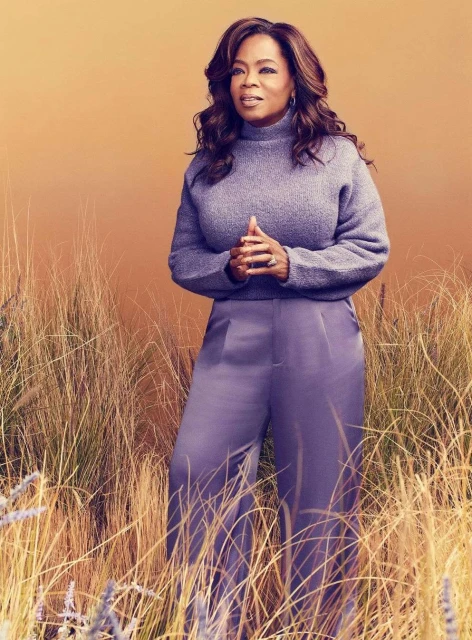 Oprah Winfrey posing in a sweater and pants in a wheat field, looking left, against a gradient orange background in a portrait shot.