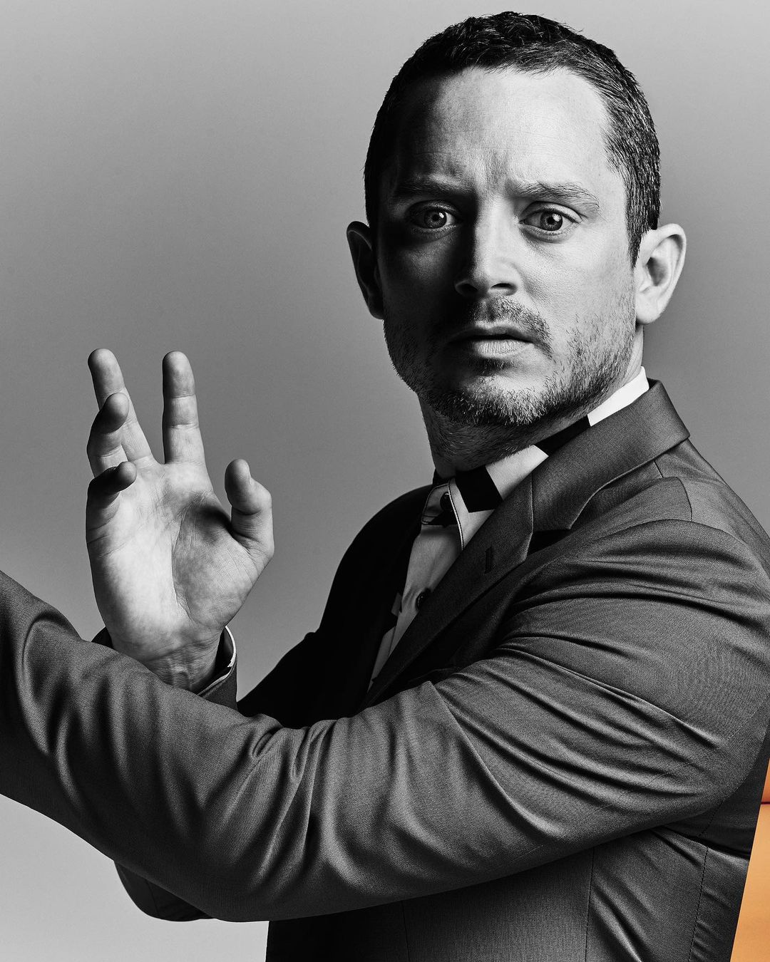 Portrait of a man in a suit and bow tie, facing the camera with an open palm gesture against a neutral gray background.