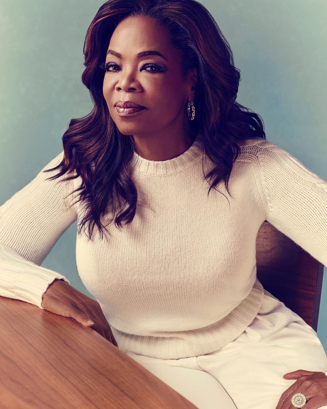 Portrait of Oprah Winfrey indoors, wearing a white sweater and pants, seated at a wooden table, with a serene expression.