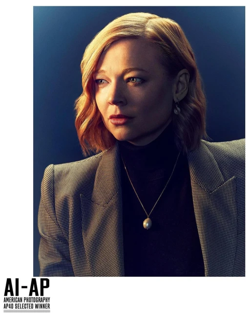 Portrait of a blonde woman in a grey blazer and black turtleneck against a dark blue backdrop, captured in a professional studio setting.