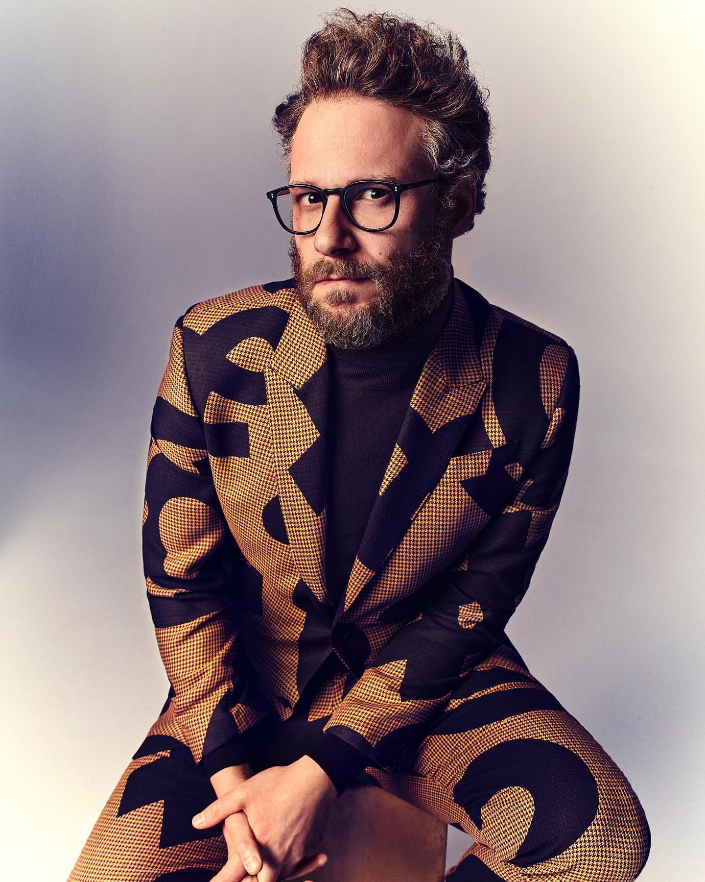 Portrait of Seth Rogen, an adult man wearing glasses and a patterned suit, seated indoors against a neutral background.