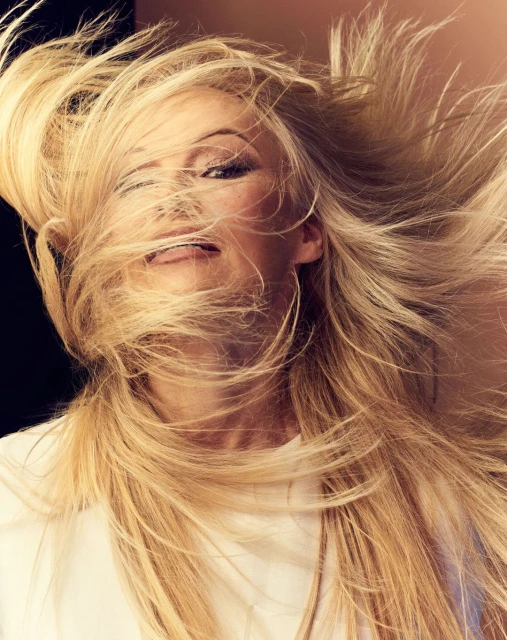 Blonde woman with flowing hair poses in a beauty shot. Her hair is tousled around her face, and she wears a white top. 