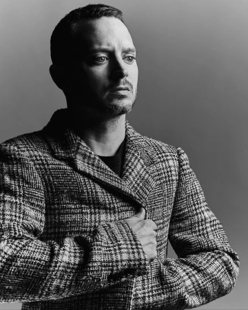 Portrait of actor Elijah Wood wearing a patterned houndstooth jacket against a plain grey studio backdrop