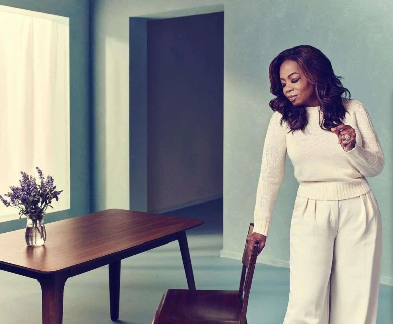 Oprah Winfrey in a light-toned room with a dark wood table with a vase of purple flowers, wearing off-white sweater and wide-leg pants.