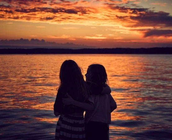 Two children embracing while standing by a lake during a vibrant sunset sky