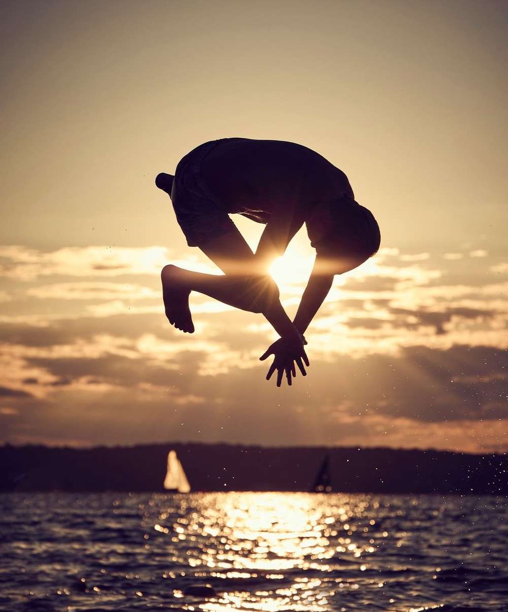 Silhouette of an adult male performing a backflip over water with a sunset sky and sailboats in the background during golden hour.