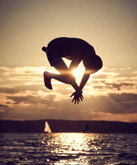Silhouette of an adult male performing a backflip over water with a sunset sky and sailboats in the background during golden hour.