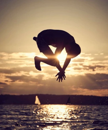 Silhouette of an adult male performing a backflip over water with a sunset sky and sailboats in the background during golden hour.