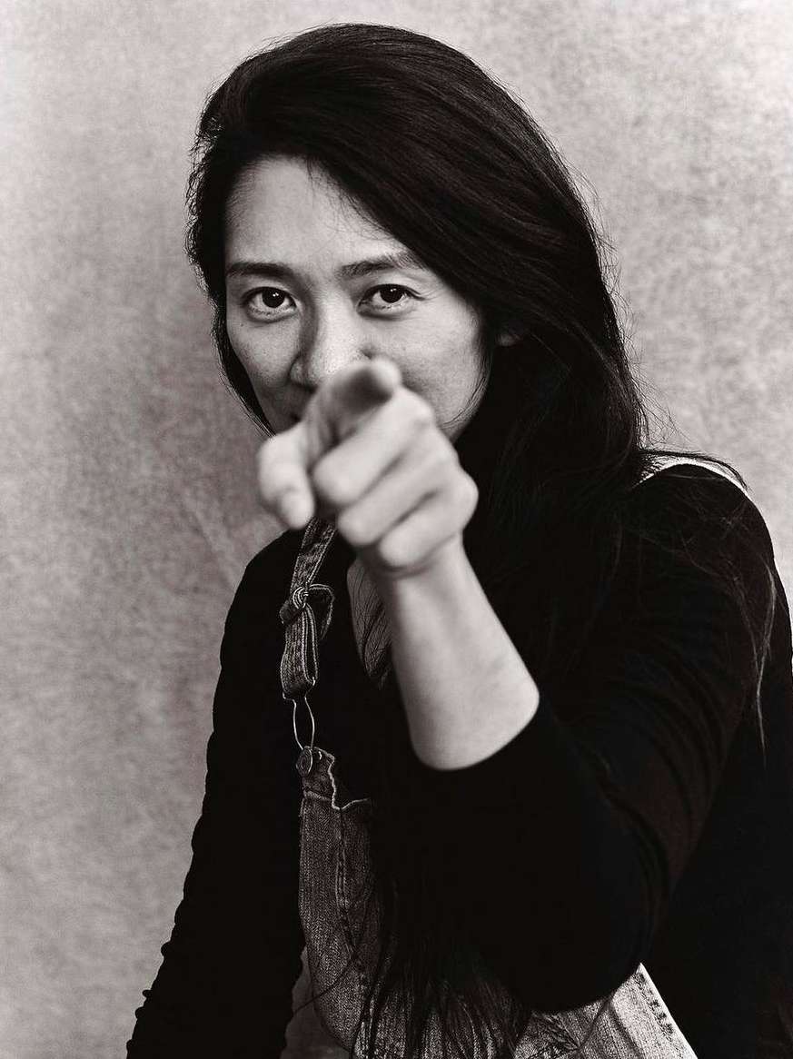 Monochrome portrait of Asian woman pointing with long black hair wearing denim overalls in a photo studio setting with a simple backdrop.
