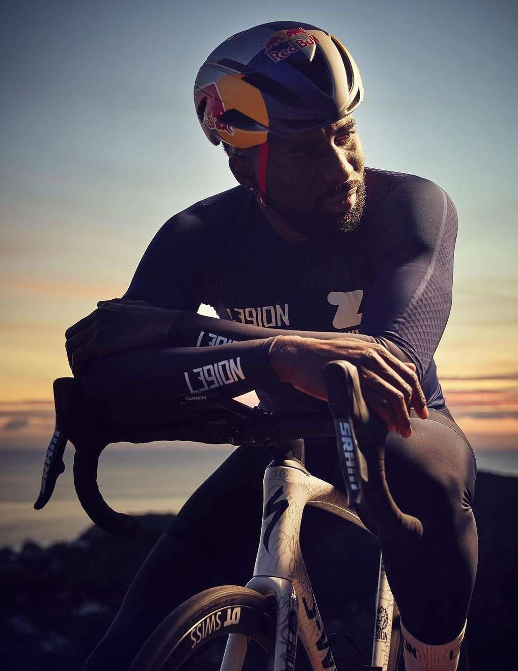 A cyclist wearing a helmet rests his arms on his bike, backlit against a sunset sky and mountain backdrop, creating a sense of athletic endurance.