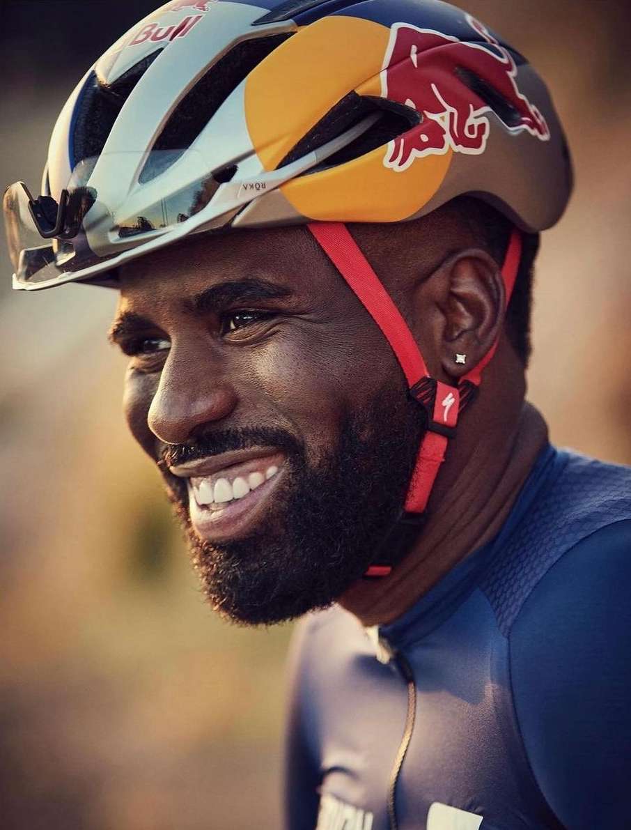 Close-up portrait of an adult male cyclist wearing a Red Bull helmet and blue cycling jersey, smiling outdoors.