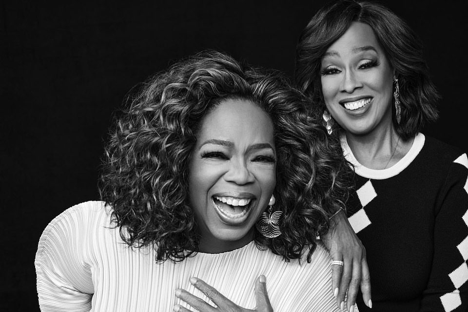 Black and white portrait of Oprah Winfrey laughing next to Gayle King against a dark background